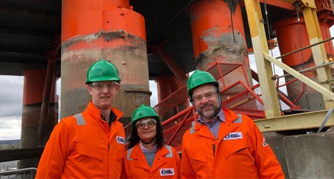 Three wokers in safety helmets and orange overalls are smiling towards the camera